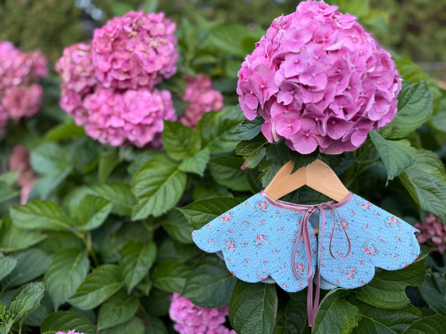 Pink hydrangeas with a blue dress collar on a hanger against a green leafy background