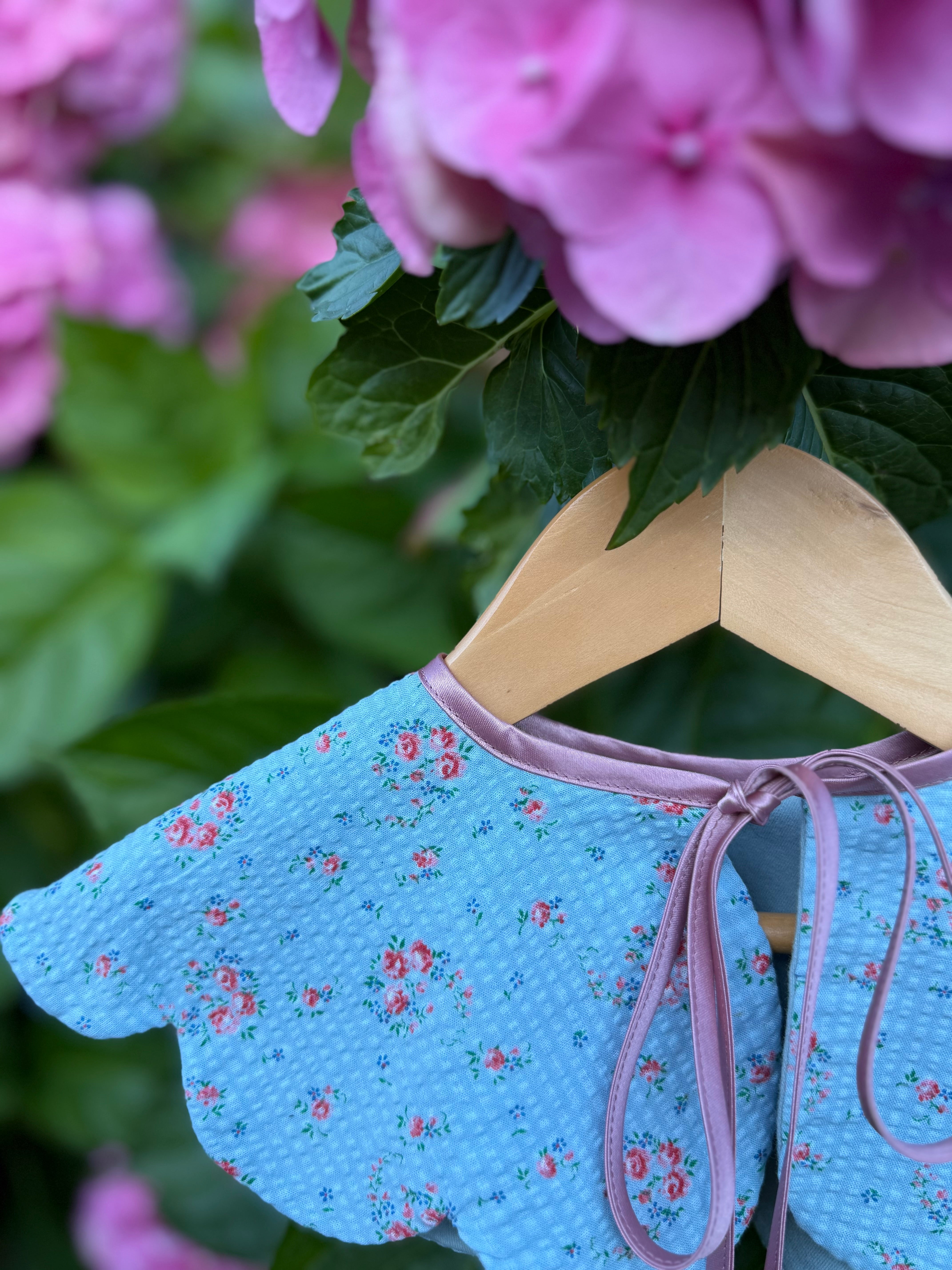 Blue floral dress  collar with pink trim on a wooden hanger against a pink flower background